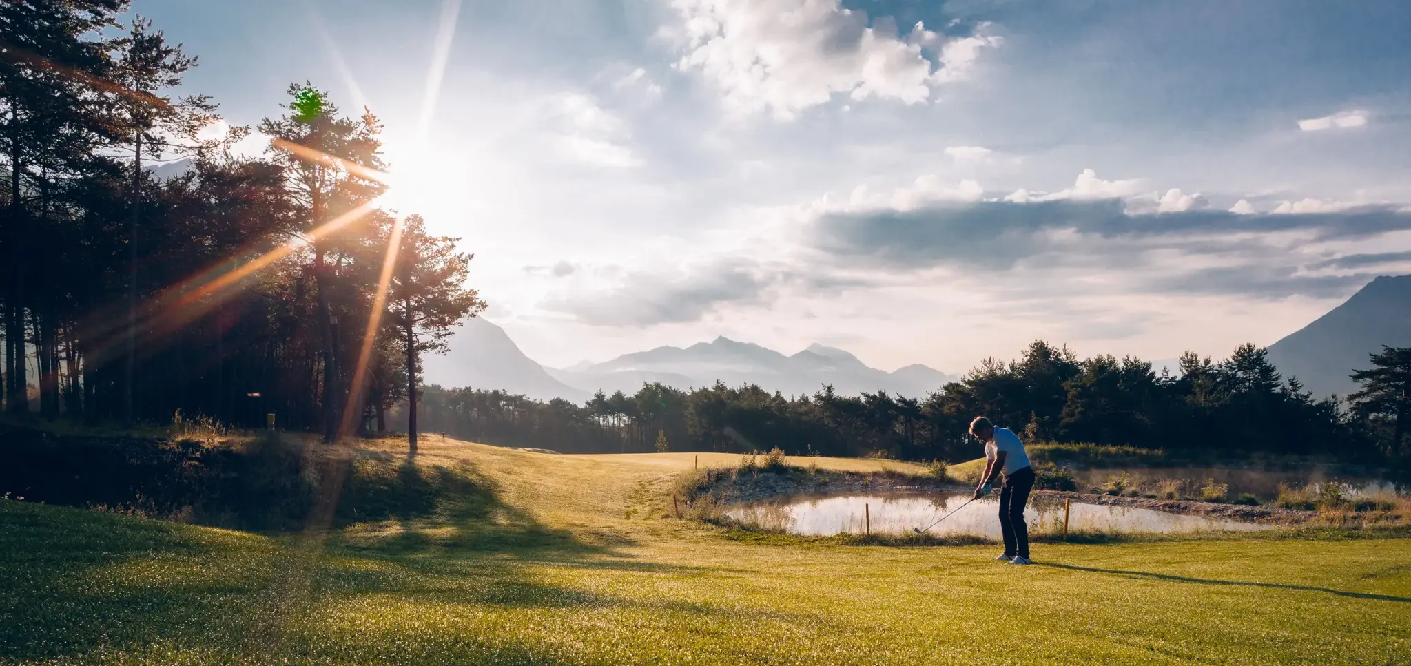 Golfer beim Abschlag auf dem Golfplatz am Mieminger Plateau, Wasserhindernis und Bergpanorama in der Abendsonne