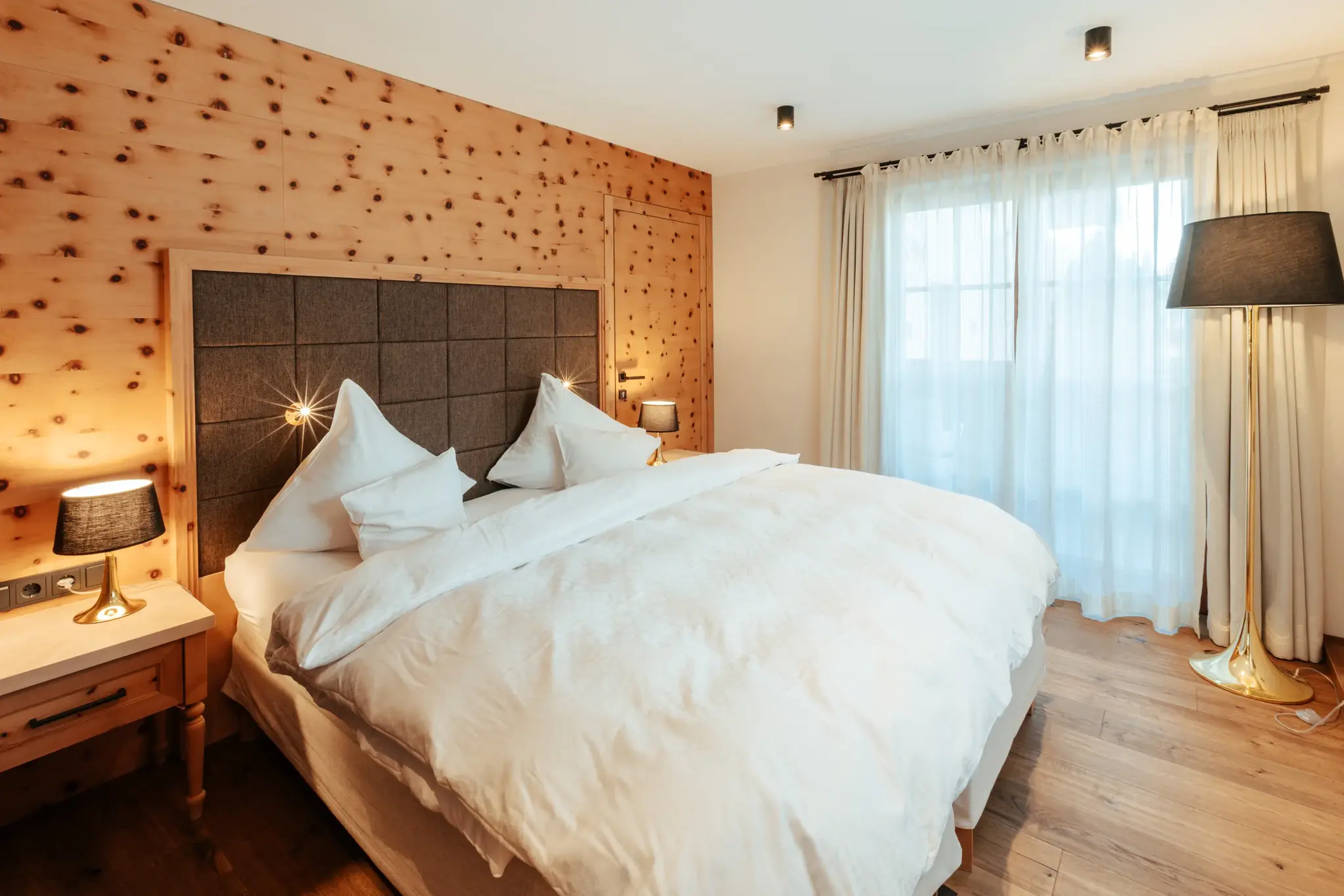 Biozimmer aus heimischer Zirbe mit gemachtem Doppelbett, warmen Nachttischlampen und hellem Fenster im Biohotel Schweitzer