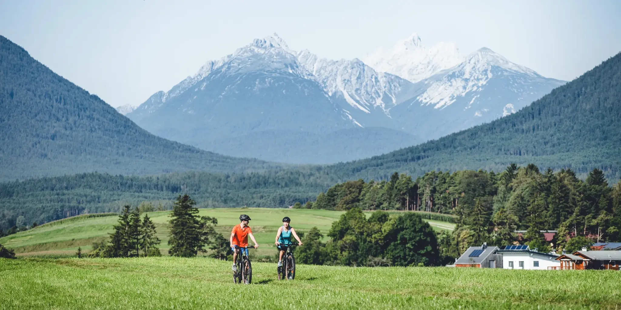 Couple mountain biking across a green summer meadow before a snow-capped mountain panorama on the Mieminger Sonnenplateau