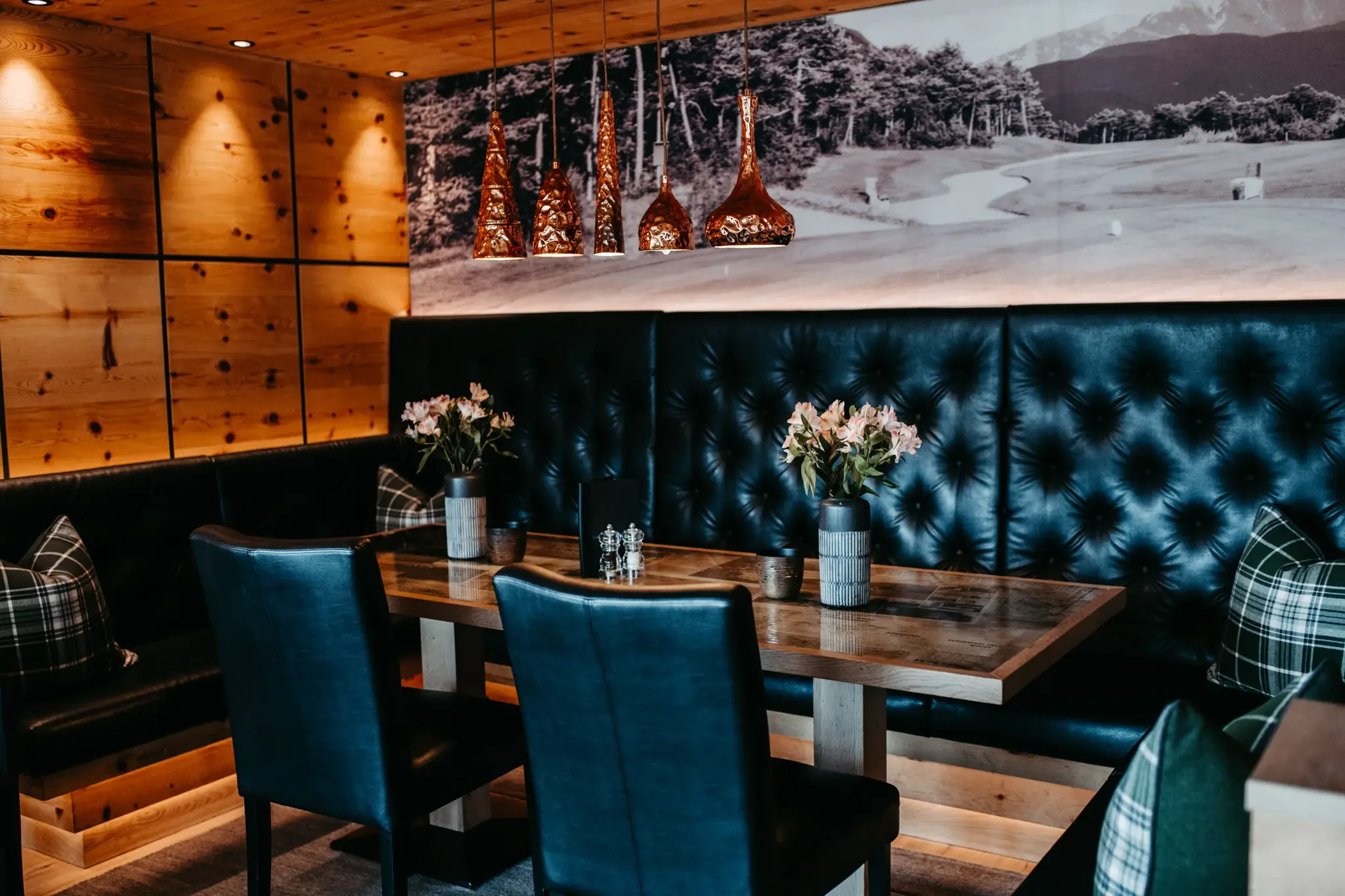Cosy seating nook at Restaurant Greenvieh with a dark leather bench, copper lights and a golf-course mural at Biohotel Schweitzer
