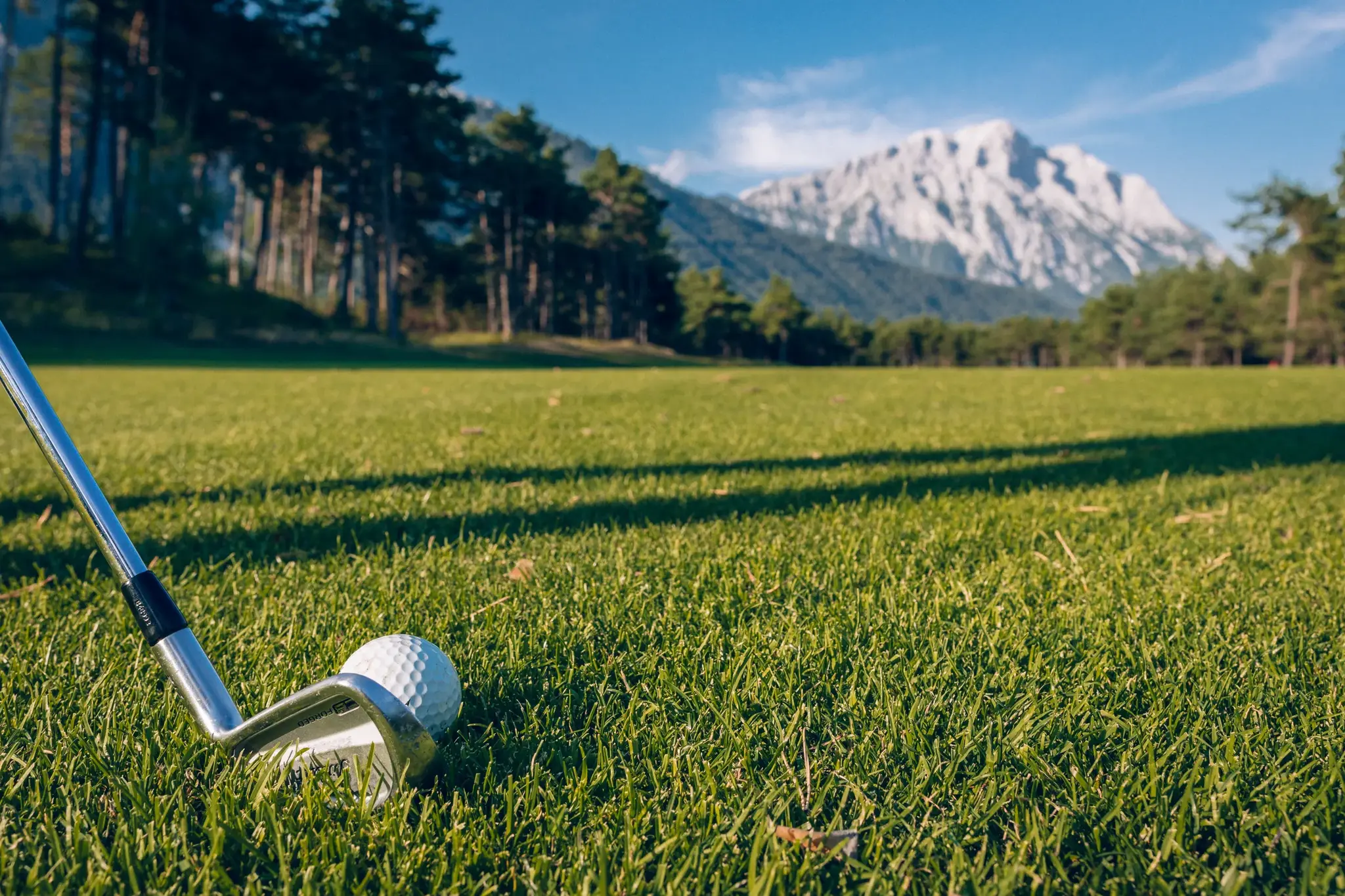 Nahaufnahme eines Golfschlägers am Ball auf dem Grün, im Hintergrund Wald und verschneite Berge am Mieminger Plateau